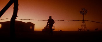 Movie still from “Razorback” (1984), directed by Russell Mulcahy – A man standing next to a barbed wire fence at sunset; Wide shot, Low angle