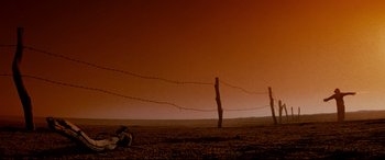 Movie still from “Razorback” (1984), directed by Russell Mulcahy – A person sitting on the ground next to barbed wire fence; Wide shot, Low angle
