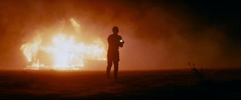 Movie still from “Razorback” (1984), directed by Russell Mulcahy – A man standing in front of a fire with a cell phone; Wide shot, Low angle