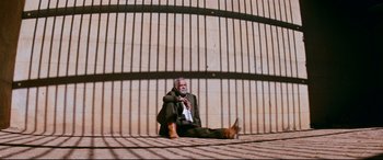 Movie still from “Razorback” (1984), directed by Russell Mulcahy – An older man sitting on the ground in front of a fence; Wide shot, Low angle