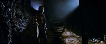 Movie still from “Razorback” (1984), directed by Russell Mulcahy – A man standing next to stairs in a dark room; Wide shot, Low angle