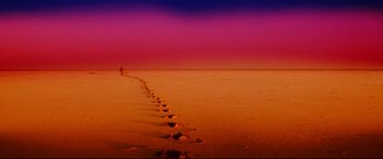 Movie still from “Razorback” (1984), directed by Russell Mulcahy – A person walking on the beach with footprints in the sand; Extreme Wide shot, High angle