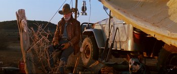 Movie still from “Razorback” (1984), directed by Russell Mulcahy – An older man in a cowboy hat sitting next to a jeep; Wide shot, Low angle