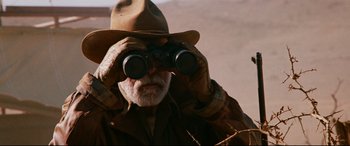 Movie still from “Razorback” (1984), directed by Russell Mulcahy – An older man wearing a hat looking through binoculars; Close Up shot, Over the shoulder angle