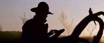Movie still from “Razorback” (1984), directed by Russell Mulcahy – A man in a hat is looking at his cell phone; Close Up shot, Low angle