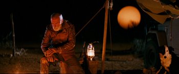 Movie still from “Razorback” (1984), directed by Russell Mulcahy – An old man in a brown jacket is lighting a lantern; Wide shot, Over the shoulder angle