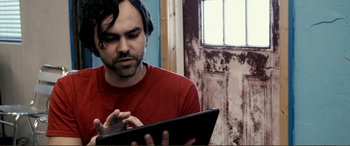 Movie still from “Hostage 911” (2018), directed by Robert Rodriguez – A man in a red shirt using a tablet; Close Up shot, Overhead angle