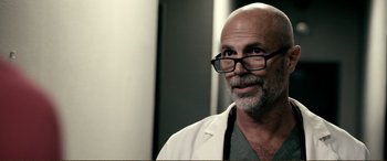 Movie still from “Hostage 911” (2018), directed by Robert Rodriguez – An older man wearing glasses and a lab coat; Close Up shot, Over the shoulder angle