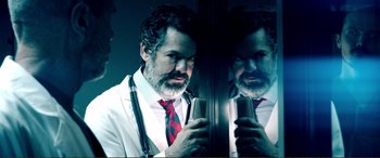 Movie still from “Hostage 911” (2018), directed by Robert Rodriguez – A man in a white lab coat holding a cell phone in front of a mirror; Medium shot, Low angle
