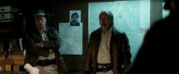 Movie still from “Red Hill” (2010), directed by Patrick Hughes – A man standing in front of a wall with a picture of a man on it; Medium shot, Over the shoulder angle