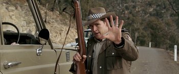 Movie still from “Red Hill” (2010), directed by Patrick Hughes – A man holding a rifle in front of a car; Close Up shot, Over the shoulder angle