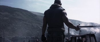 Movie still from “Reign of Fire” (2002), directed by Rob Bowman – A man in a leather vest is standing in front of a hill; Medium shot, Over the shoulder angle