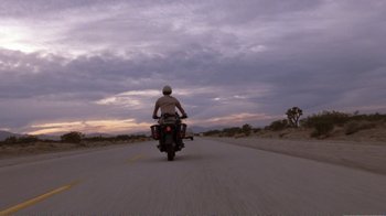 Movie still from “Repo Man” (1984), directed by Alex Cox – A man riding a motorcycle down the middle of a road; Wide shot, Low angle