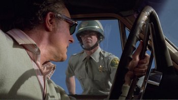 Movie still from “Repo Man” (1984), directed by Alex Cox – A man in a police uniform holding a gun in a car; Medium shot, Over the shoulder angle
