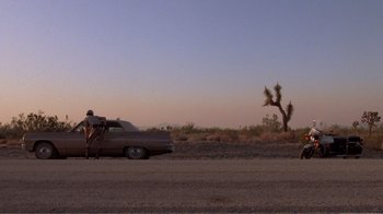 Movie still from “Repo Man” (1984), directed by Alex Cox – A man standing on the side of a road next to a car; Extreme Wide shot, Low angle