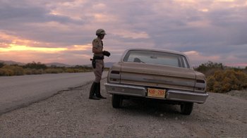 Movie still from “Repo Man” (1984), directed by Alex Cox – A police officer standing on the side of the road next to a car; Wide shot, Low angle
