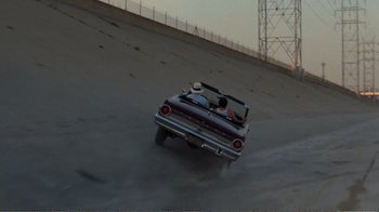 Movie still from “Repo Man” (1984), directed by Alex Cox – Two people in a convertible driving down a dirt road; Wide shot, High angle