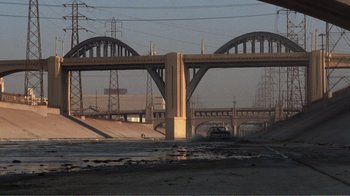 Movie still from “Repo Man” (1984), directed by Alex Cox – A car driving on a road under a bridge; Extreme Wide shot, High angle