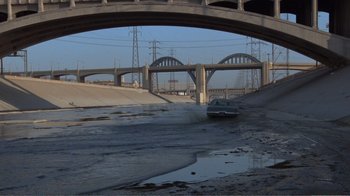 Movie still from “Repo Man” (1984), directed by Alex Cox – A car driving under a bridge over a river; Extreme Wide shot, High angle