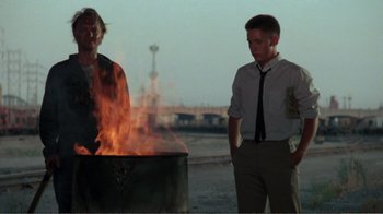 Movie still from “Repo Man” (1984), directed by Alex Cox – Two men standing in front of a fire pit; Medium shot, Over the shoulder angle