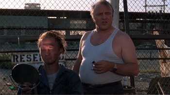 Movie still from “Repo Man” (1984), directed by Alex Cox – Two men standing next to each other in front of a chain link fence; Medium shot, Low angle