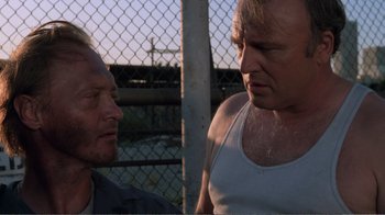 Movie still from “Repo Man” (1984), directed by Alex Cox – Two men standing next to each other in front of a chain link fence; Close Up shot, Low angle
