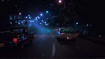 Movie still from “Repo Man” (1984), directed by Alex Cox – Cars driving down a street at night in the dark; Wide shot, High angle