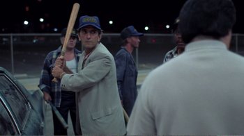 Movie still from “Repo Man” (1984), directed by Alex Cox – A man holding a baseball bat while standing next to a group of people; Medium shot, Over the shoulder angle