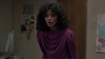 Movie still from “Repo Man” (1984), directed by Alex Cox – A woman in a purple dress is standing in front of a wall; Close Up shot, Over the shoulder angle