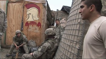Movie still from “Restrepo” (2010), directed by Tim Hetherington – A group of men in military fatigues standing next to each other; Wide shot, Over the shoulder angle