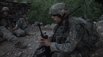 Movie still from “Restrepo” (2010), directed by Tim Hetherington – A man in a military uniform sitting on the ground holding a rifle; Medium shot, High angle