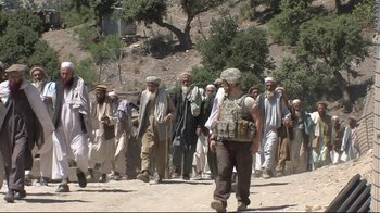 Movie still from “Restrepo” (2010), directed by Tim Hetherington – A group of men standing next to each other on a dirt road; Wide shot, Over the shoulder angle