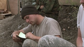 Movie still from “Restrepo” (2010), directed by Tim Hetherington – A man writing on a piece of paper while squatting on the ground; Close Up shot, High angle
