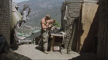 Movie still from “Restrepo” (2010), directed by Tim Hetherington – A shirtless man standing in front of a table; Wide shot, Over the shoulder angle