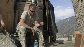 Movie still from “Restrepo” (2010), directed by Tim Hetherington – A man sitting on the side of a building next to a bottle of water; Medium shot, Low angle