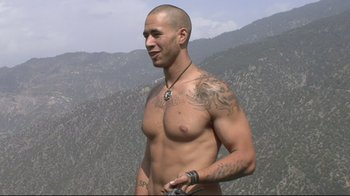 Movie still from “Restrepo” (2010), directed by Tim Hetherington – A man with tattoos is standing in front of a mountain; Medium shot, Low angle