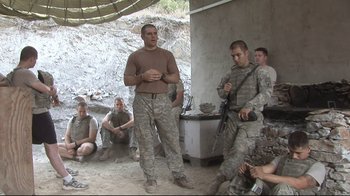 Movie still from “Restrepo” (2010), directed by Tim Hetherington – A group of men in military fatigues are sitting and standing; Wide shot, Over the shoulder angle