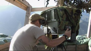 Movie still from “Restrepo” (2010), directed by Tim Hetherington – A man in a tan shirt and a hat working on a machine; Medium shot, Over the shoulder angle