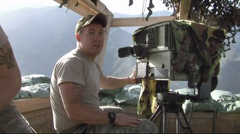 Movie still from “Restrepo” (2010), directed by Tim Hetherington – A man is sitting in a chair and holding a camera; Medium shot, High angle