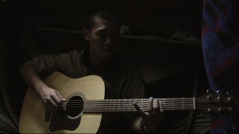 Movie still from “Restrepo” (2010), directed by Tim Hetherington – A man is playing a guitar in a dark room; Close Up shot, Low angle