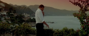 Movie still from “Revenge” (1990), directed by Tony Scott – A man standing on a hill near a body of water; Medium shot, Low angle