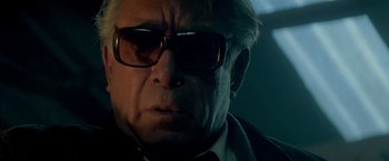 Movie still from “Revenge” (1990), directed by Tony Scott – An older man wearing glasses looking at the camera; Close Up shot, Low angle