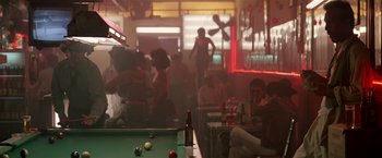 Movie still from “Revenge” (1990), directed by Tony Scott – A group of people are gathered around a pool table in a bar; Wide shot, Over the shoulder angle