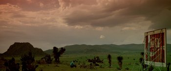 Movie still from “Revenge” (1990), directed by Tony Scott – A person sitting in a grassy field under a cloudy sky; Extreme Wide shot, Low angle