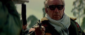 Movie still from “Revenge” (1990), directed by Tony Scott – A man holding a gun while wearing sunglasses and a scarf; Close Up shot, Low angle
