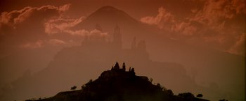 Movie still from “Revenge” (1990), directed by Tony Scott – A castle on top of a hill in front of a mountain; Extreme Wide shot, Low angle