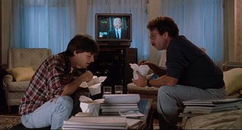 Movie still from “Reversal of Fortune” (1990), directed by Barbet Schroeder – Two men sitting in front of a television eating; Medium shot, Over the shoulder angle