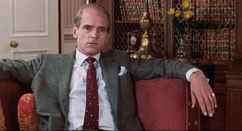 Movie still from “Reversal of Fortune” (1990), directed by Barbet Schroeder – A man in a suit and tie sitting on top of a couch; Close Up shot, Over the shoulder angle