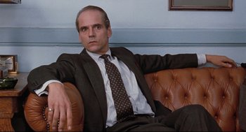 Movie still from “Reversal of Fortune” (1990), directed by Barbet Schroeder – A man in a suit and tie sitting on a couch; Medium shot, Low angle