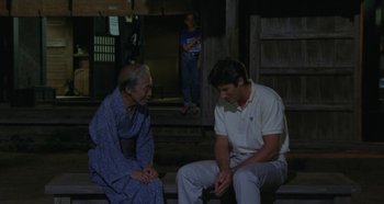 Movie still from “Rhapsody in August” (1991), directed by Akira Kurosawa – A man and a woman sitting on a porch with a child standing in the background; Wide shot, Over the shoulder angle